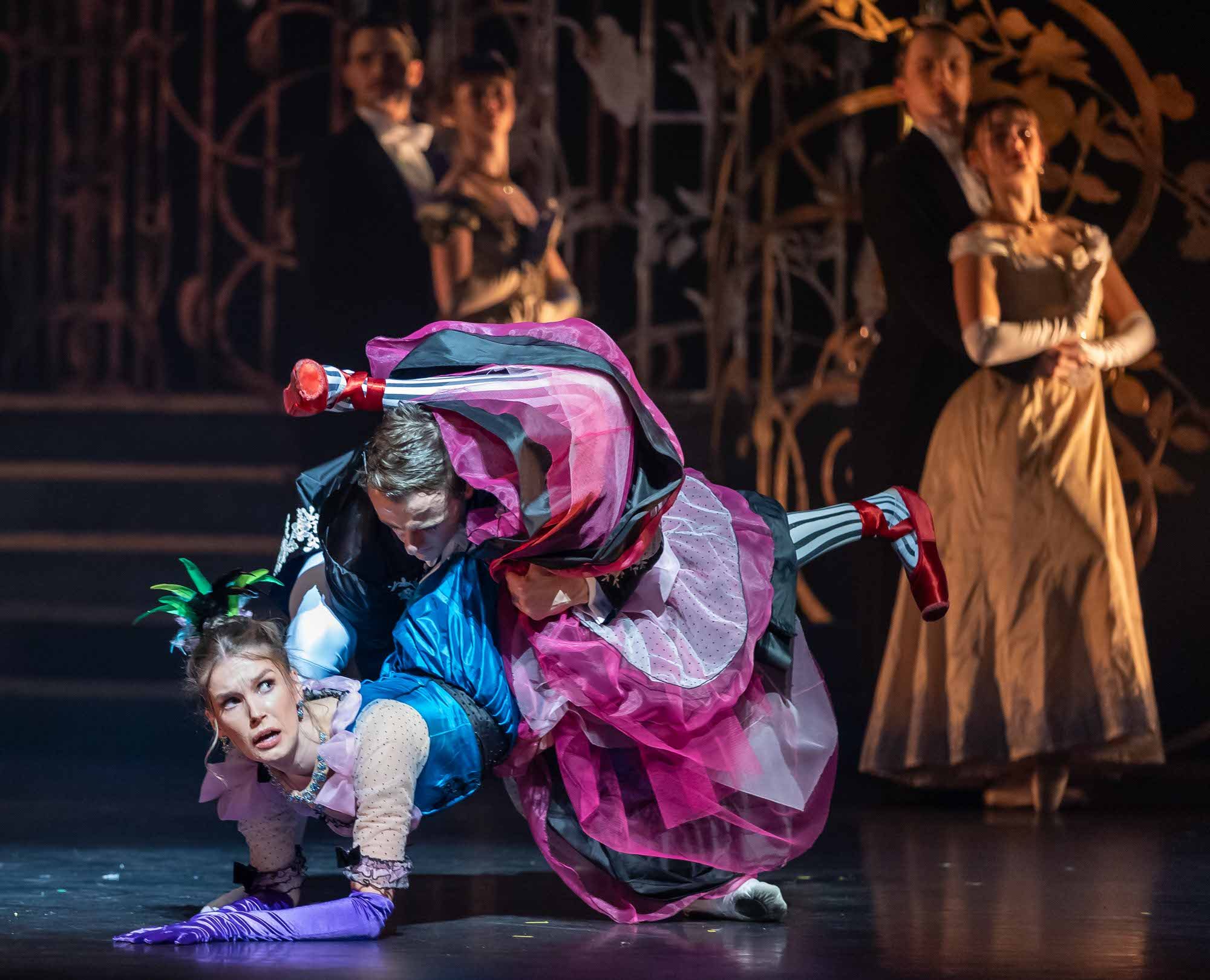 A woman in a bright, layered costume is dramatically dipped and twisted by a male dance partner on stage, while four dancers in period clothing watch in the background.